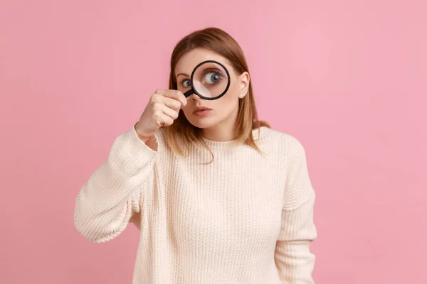 Portrait of funny blond woman standing, holding magnifying glass and looking at camera with big zoom eye, verifying authenticity, wearing white sweater. Indoor studio shot isolated on pink background.