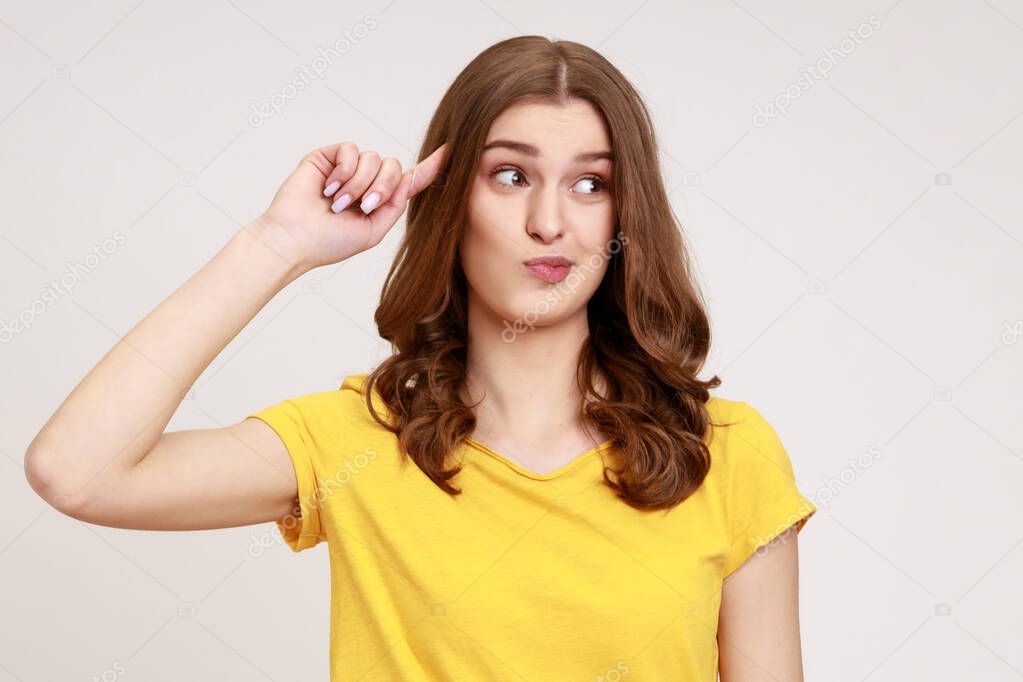 Crazy idea! Woman of young age in yellow casual T- shirt showing stupid gesture, looking at camera with condemnation and blaming for insane plan. Indoor studio shot isolated on gray background.