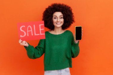 Satisfied smiling woman with Afro hairstyle wearing green casual style sweater holding sale card and smart phone with blank black display. Indoor studio shot isolated on orange background.