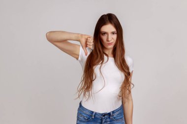 Portrait of dark haired unhappy unsatisfied woman showing thumb down, dislike gesture, negative emotions, bad rating, wearing white T-shirt. Indoor studio shot isolated on gray background.