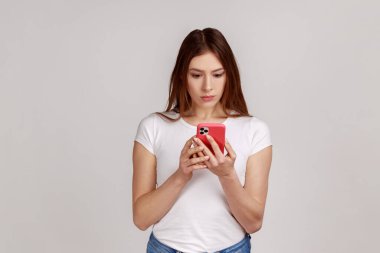 Portrait of concentrated woman using smart phone, typing message or checking social networks, writing post, serious expression, wearing white T-shirt. Indoor studio shot isolated on gray background.