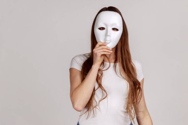 Portrait of anonymous dark haired woman covering her face with white mask, hiding personality, conspiracy and privacy, wearing white T-shirt. Indoor studio shot isolated on gray background.