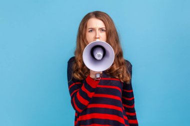 Portrait of woman wearing striped casual style sweater, taking in megaphone, looking at camera with serious expression, important announcement. Indoor studio shot isolated on blue background.