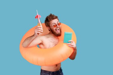 Portrait of bearded man standing with rubber ring, holding passport document and raised arm with cocktail, celebrating vacation, travel tour. Indoor studio shot isolated on blue background.