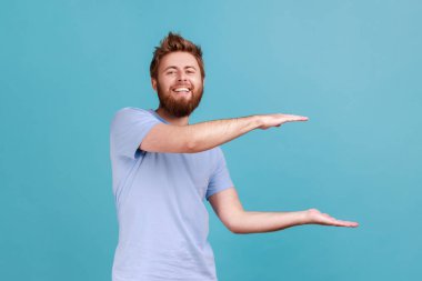 Portrait of young adult positive optimistic bearded man presenting area for advertisement, looking at camera with toothy smile and optimism. Indoor studio shot isolated on blue background.