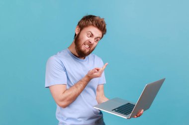 Portrait of bearded man showing middle finger to laptop screen, aggressively communicating on video call, expressing hate to job or coworker. Indoor studio shot isolated on blue background.