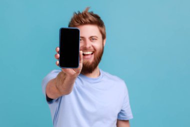 Portrait of bearded man holding out mobile phone and looking at camera with toothy smile and positive expression, being glad about technology. Indoor studio shot isolated on blue background.