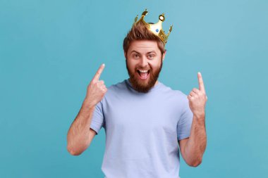Portrait of excited amazed bearded man pointing fingers on golden crown on his head, showing her leadership qualities, concept of authority. Indoor studio shot isolated on blue background. clipart