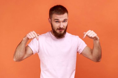 Portrait of man pointing fingers down paying attention at advertising area, showing freespace, looking at camera, wearing pink T-shirt. Indoor studio shot isolated on orange background.