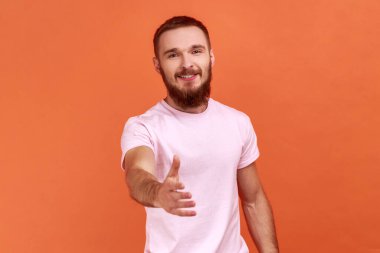 Portrait of bearded man standing with outstretched hand, offering handshake to partner, greeting on job interview, wearing pink T-shirt. Indoor studio shot isolated on orange background.