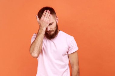 Portrait of bearded man holding big clock and biting nails, nervous about delayed meeting or deadline, need hurry up, wearing pink T-shirt. Indoor studio shot isolated on orange background.