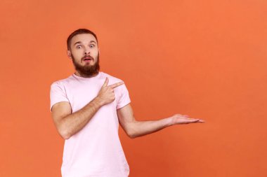 Portrait of surprised bearded man pointing finger right at empty copy space with astonishment, place for advertisement, wearing pink T-shirt. Indoor studio shot isolated on orange background.