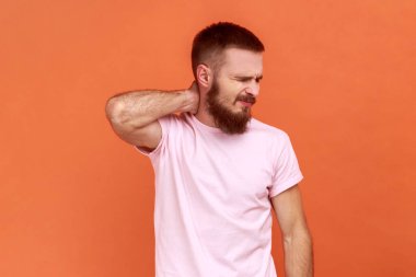 Portrait of sick bearded man grimaces from intense pain, massages neck to reveal muscle tension, injured backbone, feels bad, wearing pink T-shirt. Indoor studio shot isolated on orange background.