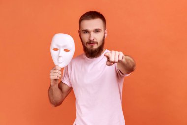 Portrait of serious bearded man pointing finger to camera and holding white mask in his hand, wants to change personality, wearing pink T-shirt. Indoor studio shot isolated on orange background.