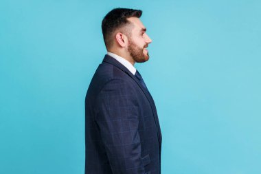 Profile portrait of young adult handsome bearded businessman wearing official style suit looking ahead with toothy smile, optimism, success. Indoor studio shot isolated on blue background.