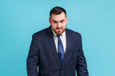Angry handsome businessman wearing official style suit looking at camera, having aggressive facial expression, expressing negative, hatred. Indoor studio shot isolated on blue background.