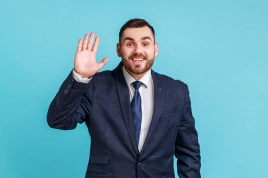 Hello! Portrait of friendly glad handsome brunette man with beard wearing official style suit showing hi gesture with waving hand and smiling sincerely Indoor studio shot isolated on blue background.