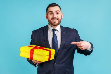 Happy man with beard wearing official style suit standing, holding yellow gift box, pointing finger, looking at camera with toothy smile. Indoor studio shot isolated on blue background.