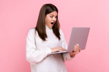Side view of astonished female working on laptop, being shocked of results of her work, open mouth, wearing white casual style sweater. Indoor studio shot isolated on pink background.