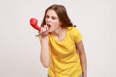 Housing maintenance call center. Angry brown haired teenager girl, shouting into retro phone handset, irritated by customer call, wears yellow T-shirt. Indoor studio shot isolated on gray background.
