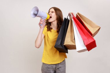 Young beautiful woman with brown wavy hair wears yellow casual style T-shirt holding shopping bags and megaphone, announcing sale and discount. Indoor studio shot isolated on gray background.