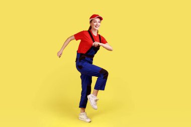 Full length portrait of optimistic positive worker woman marching, posing on one leg, going to do her work, wearing overalls and red cap. Indoor studio shot isolated on yellow background.