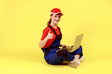 Side view portrait of optimistic worker woman sitting on floor with crossed legs and working on notebook, showing thumb up, wearing overalls and cap. Indoor studio shot isolated on yellow background.