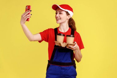Attractive delivery woman with toothy smile holding ordered coffee and having video call with client, wearing overalls and red cap. Indoor studio shot isolated on yellow background.