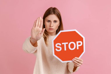 Portrait of serious woman holding red stop sign, looking at camera with ban palm gesture, has strict expression, prohibition, wearing white sweater. Indoor studio shot isolated on pink background.