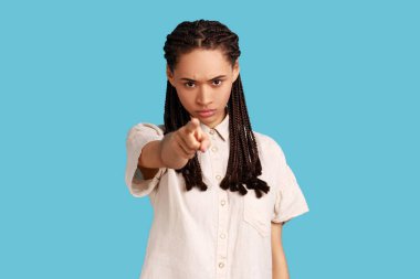 Serious strict woman with black dreadlocks looking attentively at camera, pointing directly at camera, choosing you, wearing white shirt. Indoor studio shot isolated on blue background.