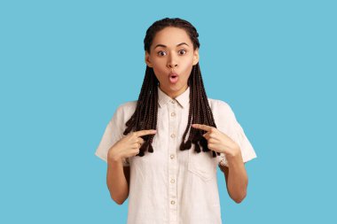 Astonished young woman with black dreadlocks pointing at herself, asks who me, has surprised expression, shocked being picked, wearing white shirt. Indoor studio shot isolated on blue background.