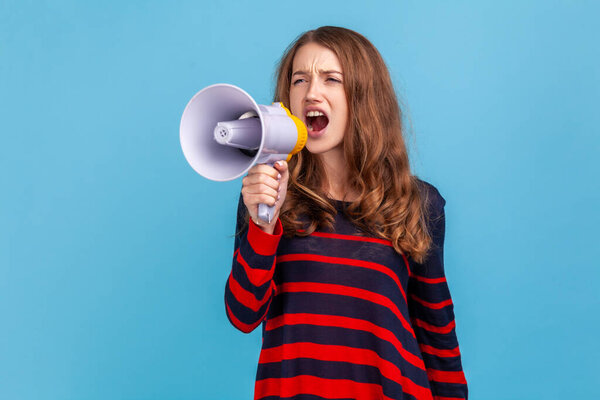 Portrait of woman wearing striped casual style sweater, screaming loudly in megaphone, announcing important advertisement, protesting. Indoor studio shot isolated on blue background.