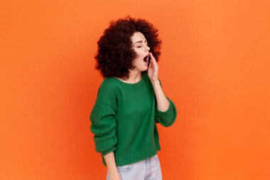 Afro saç stili, yeşil günlük süveter giyen, esneyen, ağzı avuç içi açık bir kadının portresi. Turuncu arka planda kapalı stüdyo çekimleri.