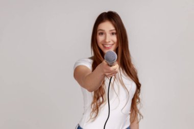 Siyah saçlı, gülümseyen, mikrofon uzatan, röportaj yapan, beyaz tişört giyen çekici bir kadının portresi. Gri arka planda izole edilmiş kapalı stüdyo görüntüleri.