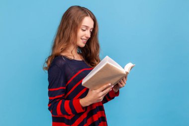 Pozitif kadının çizgili, sıradan bir süveter giymesi, okumayı sevmesi, heyecan verici bir hikayeyle ayakta durması, planlama programını kontrol etmesi. Mavi arka planda kapalı stüdyo çekimi