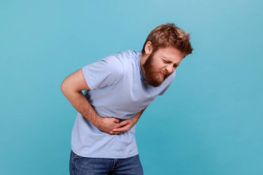 Mide ağrısı. Sakallı adamın ayakta ve acı veren karnını tutarken kötü hissederken, ishal ya da kabızlık çekerken, apandisit riski altındayken. Mavi arka planda kapalı stüdyo çekimi.