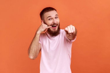 Kulağına yakın bir yerde sakallı bir adamın portresi telefon hareketlerini gösteriyor kameraya bakıyor ve senin aramanı bekliyor, pembe tişört giyiyor. Turuncu arka planda kapalı stüdyo çekimleri