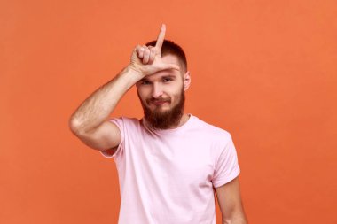 Alnının yakınında el ele tutuşan sakallı bir adamın portresi parmaklarıyla ezik jestler gösteriyor, finansal ve iş sorunları yaşıyor, pembe tişört giyiyor. Turuncu arka planda kapalı stüdyo çekimleri.