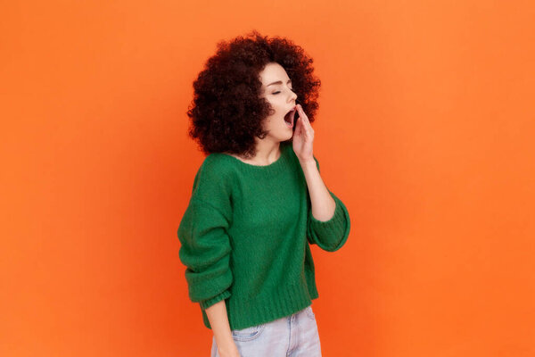 Profile portrait of sleepy woman with Afro hairstyle wearing green casual style sweater standing yawning, covering open mouth with palm. Indoor studio shot isolated on orange background.