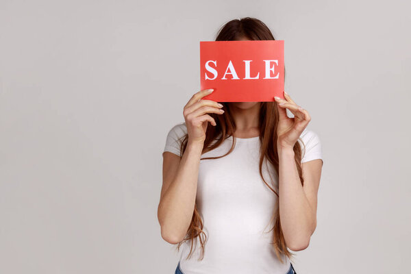 Portrait of anonymous unknown young woman hiding her face behind card with sale inscriptions, shopping, discounts, wearing white T-shirt. Indoor studio shot isolated on gray background.
