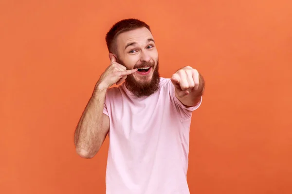 大胡子男人的画像 他的手指紧握着耳朵 显示出电话手势 看着相机 等待你的电话 穿着粉色T恤 在橙色背景下被隔离的室内拍摄 — 图库照片