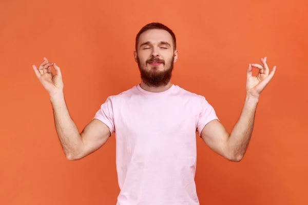 一个留着胡子的男人的画像 他高举双臂站在那里 做瑜伽冥想运动 做泥巴手势 穿着粉色T恤 在橙色背景下被隔离的室内拍摄 — 图库照片
