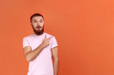 Şaşırmış sakallı adamın portresi, kameraya bakıyor, şaşırmış bir yüz ve duvara bakıyor, pembe tişört giyiyor. Turuncu arka planda kapalı stüdyo çekimleri.