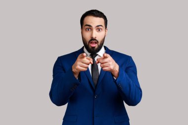 Şaşırmış sakallı adam kamerayı işaret ediyor, yüzünde şok olmuş bir ifadeyle duruyor, inanması zor, resmi stil takım elbise giyiyor. Gri arka planda izole edilmiş kapalı stüdyo görüntüleri.