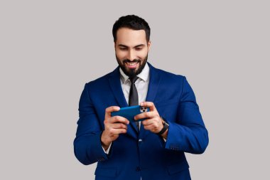Mutlu bir ifadeyle cep telefonu kullanan, cep telefonuyla video oyunu oynayan, resmi stil kıyafeti giyen sakallı bir adam. Gri arka planda izole edilmiş kapalı stüdyo görüntüleri.