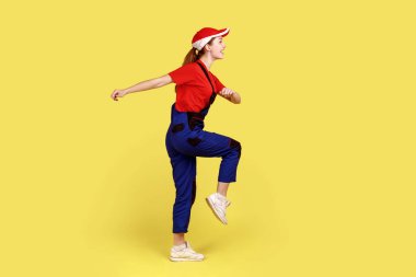 İşçi kadın yürüyüşünün, işe gidişinin, olumlu duyguları ve mutluluğu ifade edişinin, tulum ve kırmızı başlık takışının yan görünüm portresi. Sarı arka planda kapalı stüdyo çekimleri.