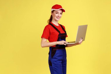 Dizüstü bilgisayarda çalışan, internetten sipariş alan, kameraya gülümseyen, tulumlu ve kırmızı şapkalı mutlu bir kadının yan görünüm portresi. Sarı arka planda kapalı stüdyo çekimleri.