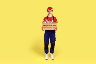 Pozitif tatmin olmuş, içi boş plastik şişelerle dolu, tulumlu ve kırmızı şapkalı bir kadın portresi. Sarı arka planda kapalı stüdyo çekimleri.