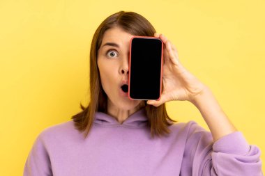 Çekici, genç, yetişkin bir kadın. Gözlerini cep telefonuyla kapatıyor. Boş bir ekran, şok edici bir ifade, mor kapüşonlu bir şey giyiyor. Sarı arka planda kapalı stüdyo çekimleri.