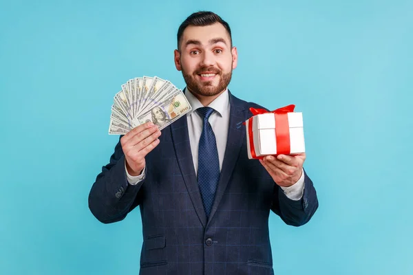 年轻英俊的男子留着胡子 身穿正式风格的西服 展示美元钞票 现金和礼品盒 看着相机 室内拍摄被蓝色背景隔离 — 图库照片